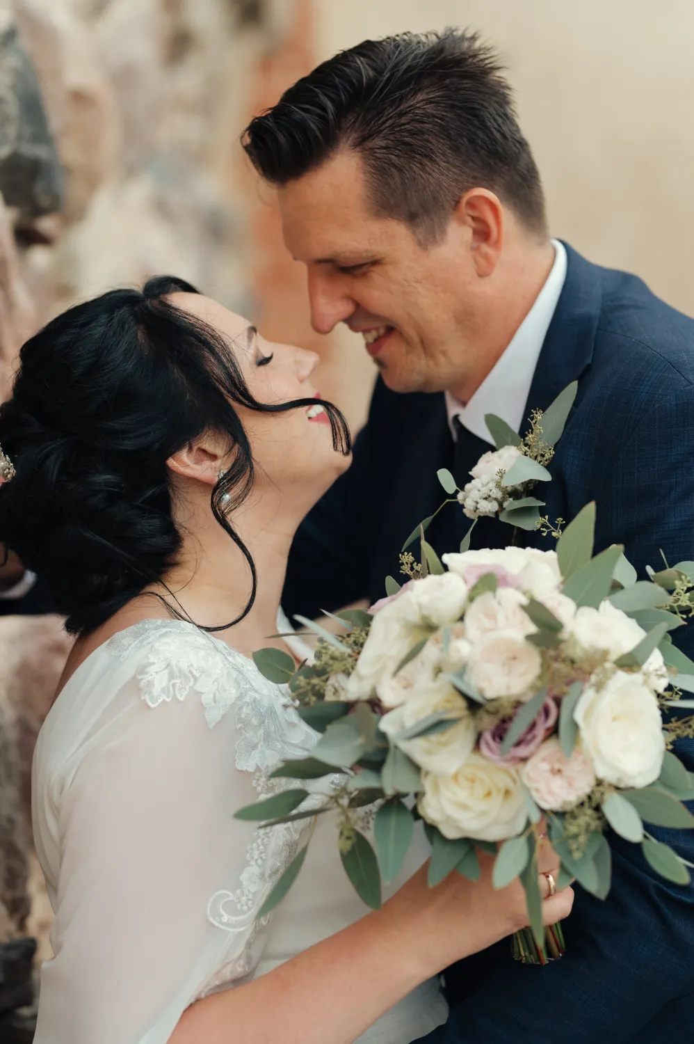 A bride in a white lace gown holds a bouquet of white and blush roses with eucalyptus, smiling as she gazes up at a groom in a dark suit who leans in close, both sharing an intimate moment against a soft, blurred stone wall.