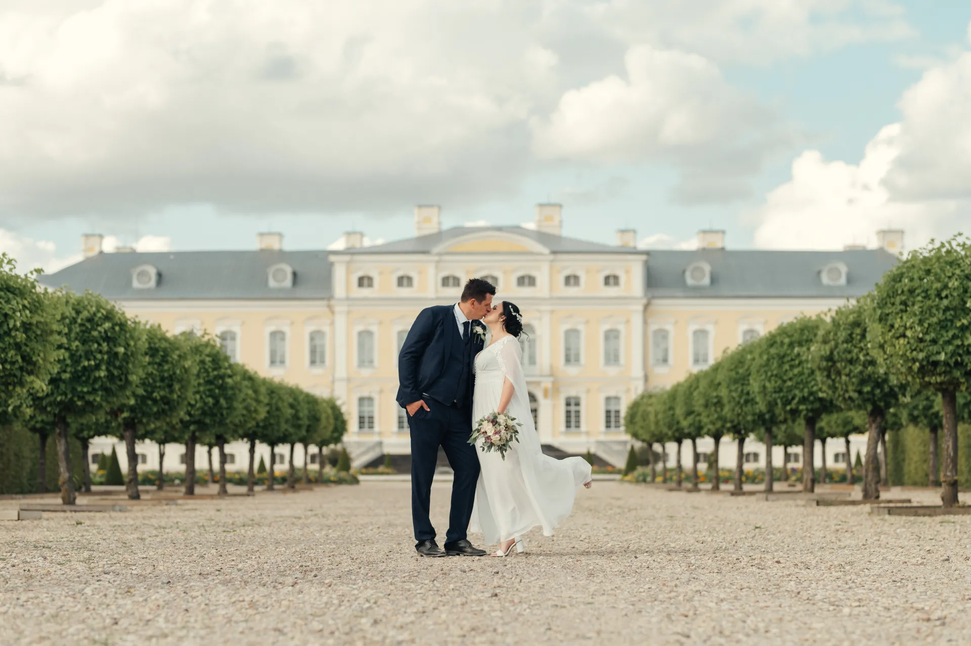 A bride in a white wedding dress holding a bouquet kisses a groom in a dark suit on a gravel courtyard in front of a large pale yellow palace with symmetrical clipped trees. The soft cloudy daylight creates a romantic wedding portrait mood.