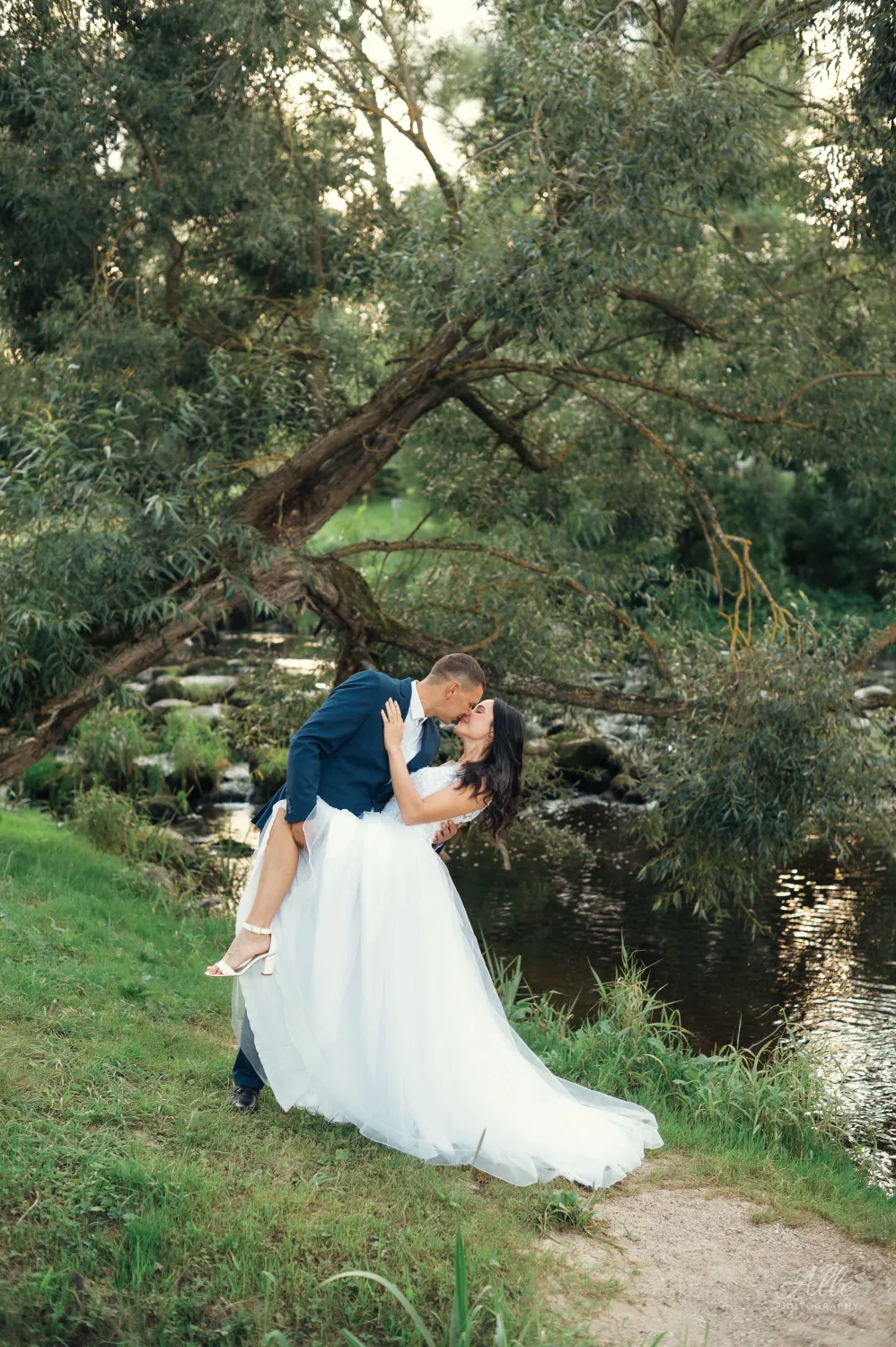A bride in a white gown and a groom in a blue suit share a kiss while he dips her on a grassy riverbank beneath leafy trees, captured in a romantic wedding portrait with soft natural light.