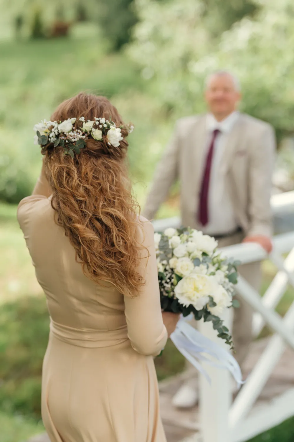 A bride with long curly hair and a floral crown holds a white bouquet, facing an older man in a beige suit and maroon tie on a white bridge in a green garden.