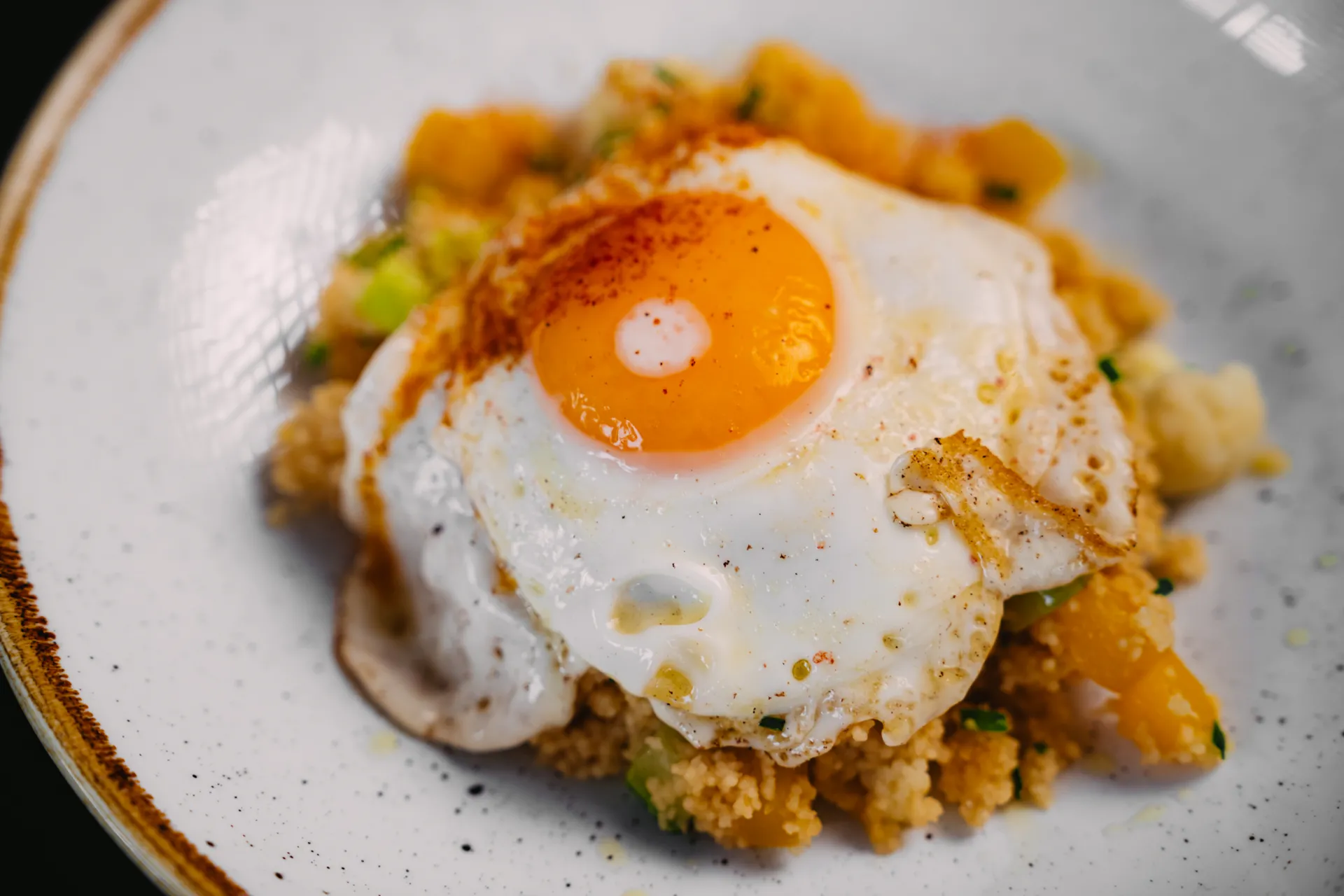 Ēdiena Fotogrāfija — Close-up food photograph of a fried egg with a bright orange yolk and a dusting of red spice resting on a bed of couscous mixed with diced orange squash and green herbs, served on a white speckled ceramic plate. The shallow depth of field and warm lighting emphasize texture in …