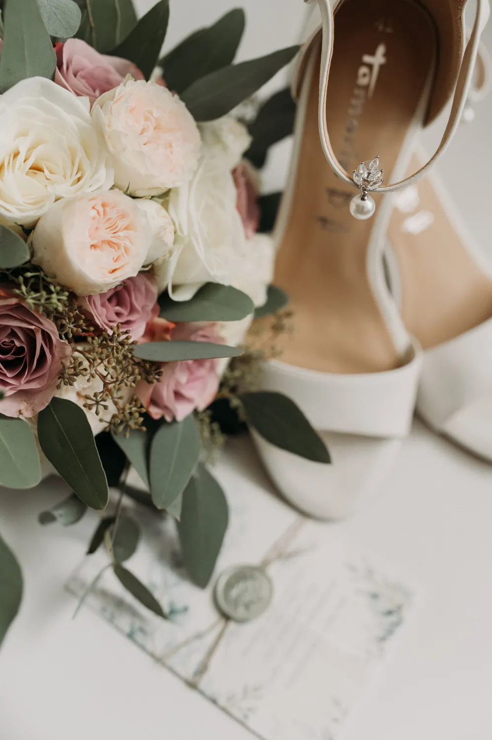 A bridal bouquet of white and blush roses with eucalyptus rests beside white strappy heels and a sealed invitation on a light surface, captured in soft natural light.