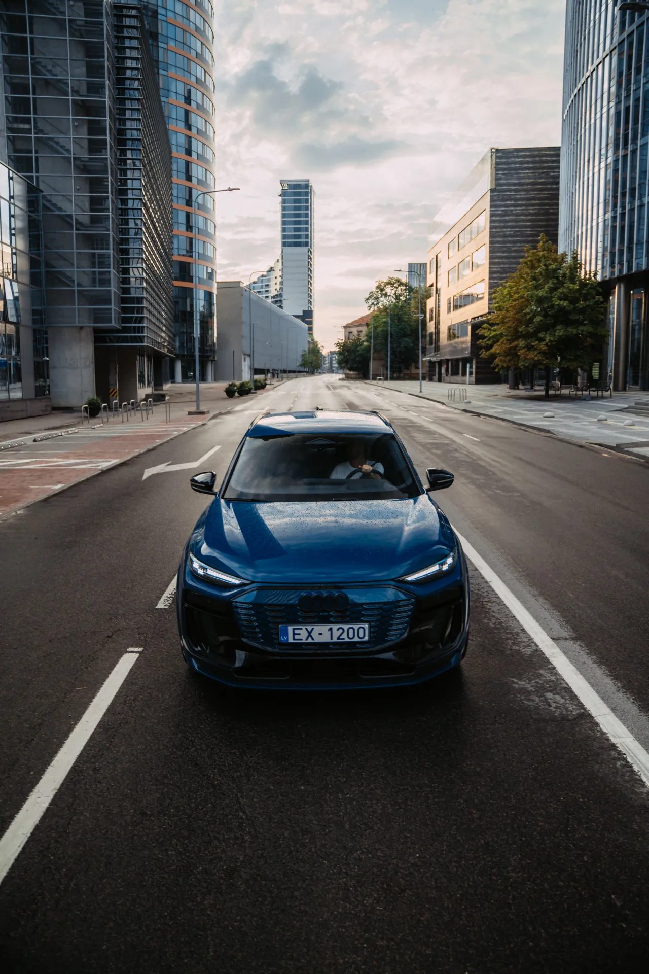 Auto Foto — A blue Audi electric SUV drives toward the camera down an empty city street lined with modern glass buildings under a cloudy sky. The driver is visible through the windshield, and the license plate reads EX-1200.