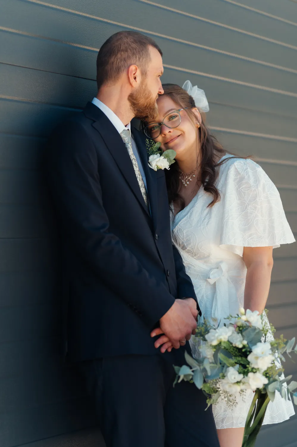 A bride in a white short dress with flutter sleeves holds a white bouquet and rests her head against the groom in a dark suit with a boutonnière, both leaning against a dark horizontal siding wall in bright daylight. Intimate wedding portrait with warm, soft natural light and a tender, relaxed mood.