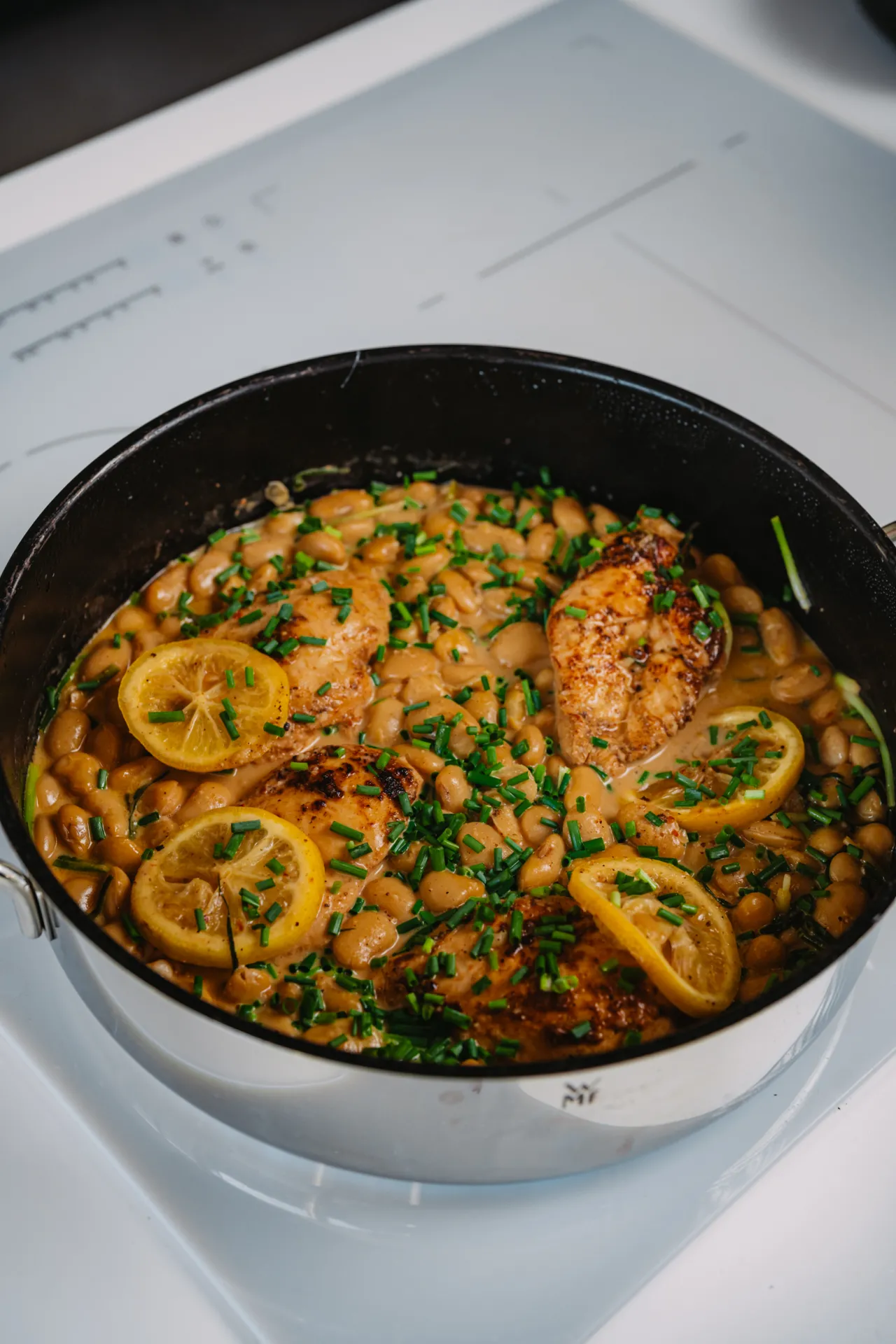 Ēdiena Fotogrāfija — A stainless steel pan with a black interior sits on a white induction cooktop, holding seared chicken breasts in a creamy white bean sauce garnished with lemon slices and chopped chives. The bright, clean overhead lighting highlights the food in a close-up commercial food photo…