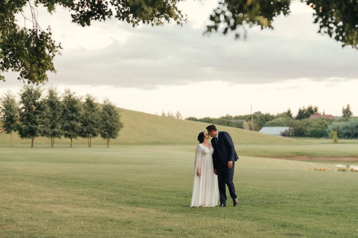 A bride in a white gown and a groom in a dark suit stand close on a grassy field, foreheads touching under tree branches, with rolling hills and distant buildings under an overcast sky in a wedding portrait.