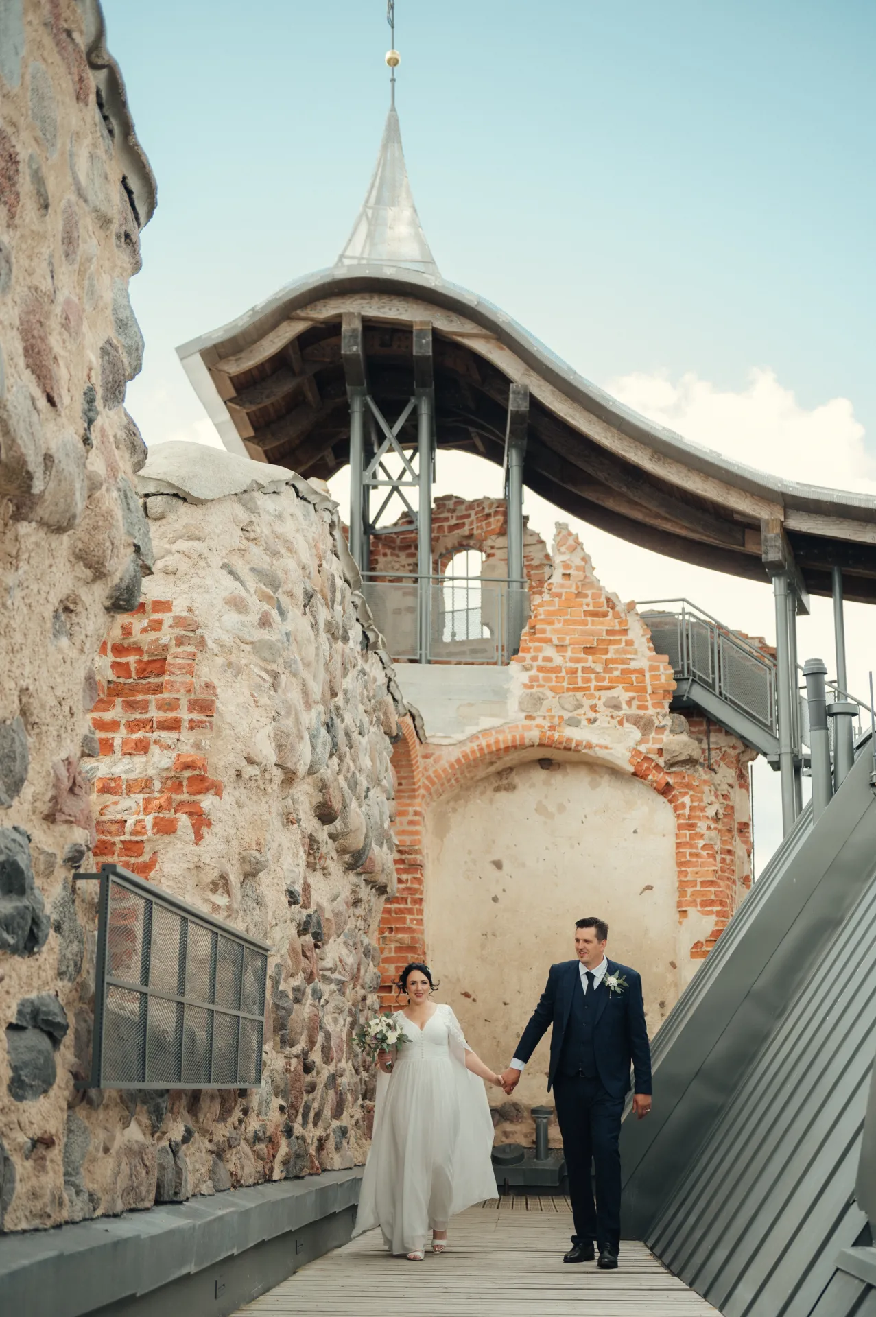 A bride in a white gown and a groom in a dark suit hold hands while walking along a wooden walkway between old stone and brick walls beneath a modern arched structure with a spire, under a bright sky.