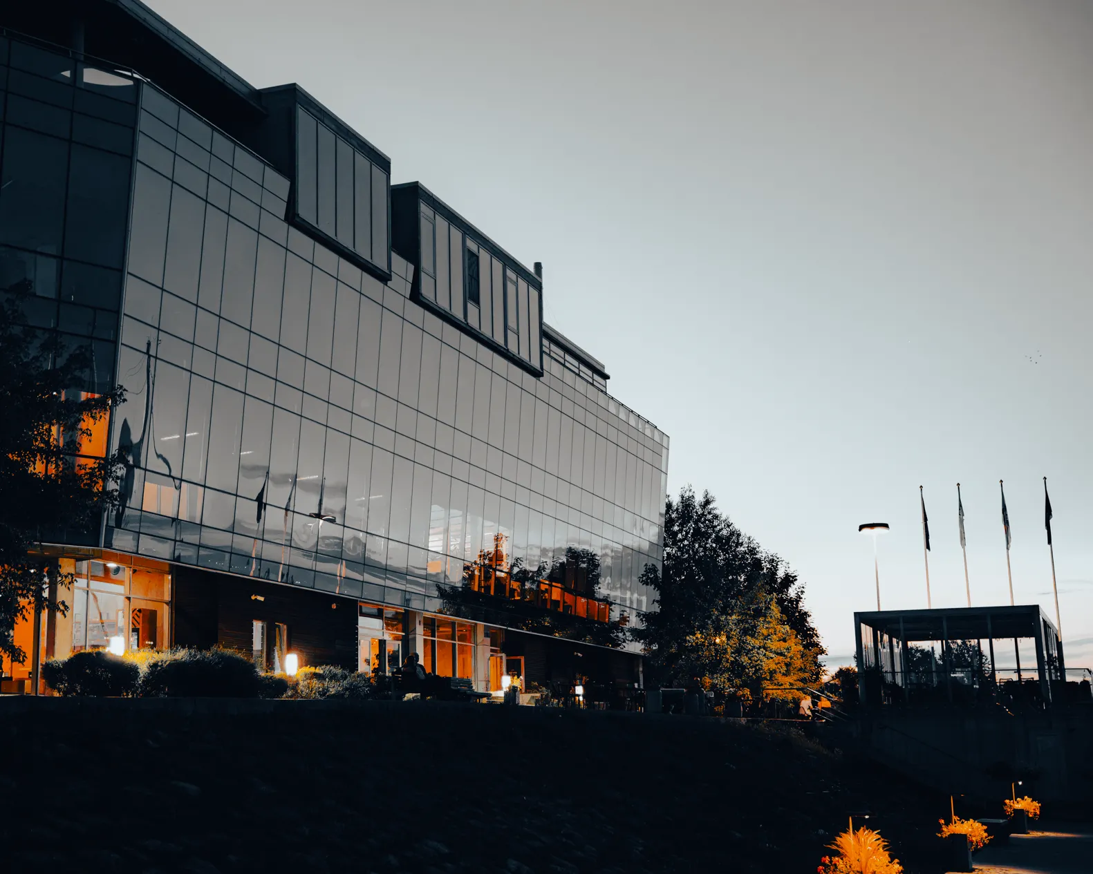 Monochrome Port View — A modern glass office building reflects the evening sky and warm interior lights, with trees and flagpoles visible beside a covered walkway under soft twilight lighting.