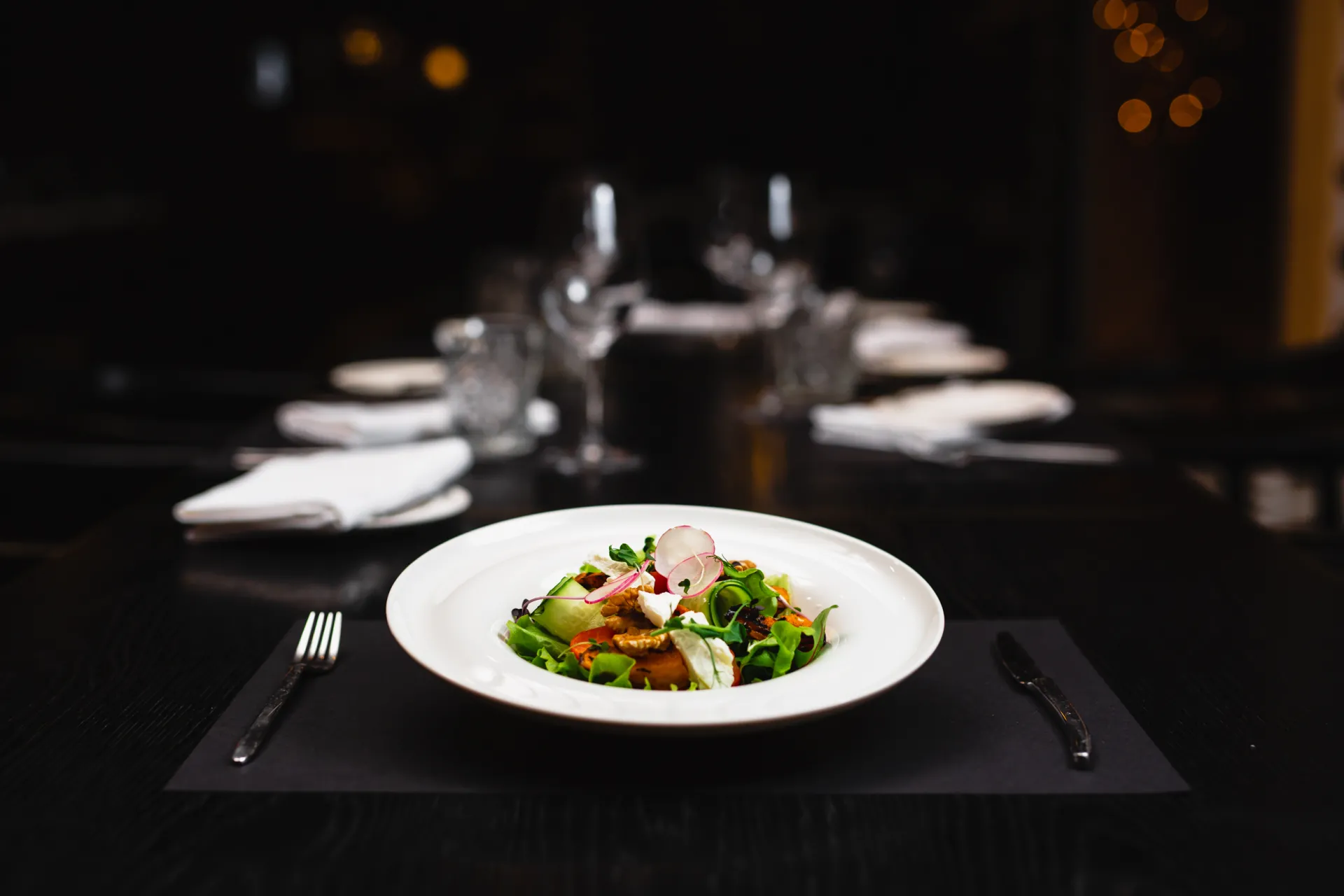 Ēdiena Fotogrāfija — A plated salad with greens, radish slices, and white cheese sits on a black placemat with silverware, set on a dark table in a dimly lit restaurant with blurred glassware and warm bokeh lights in the background.