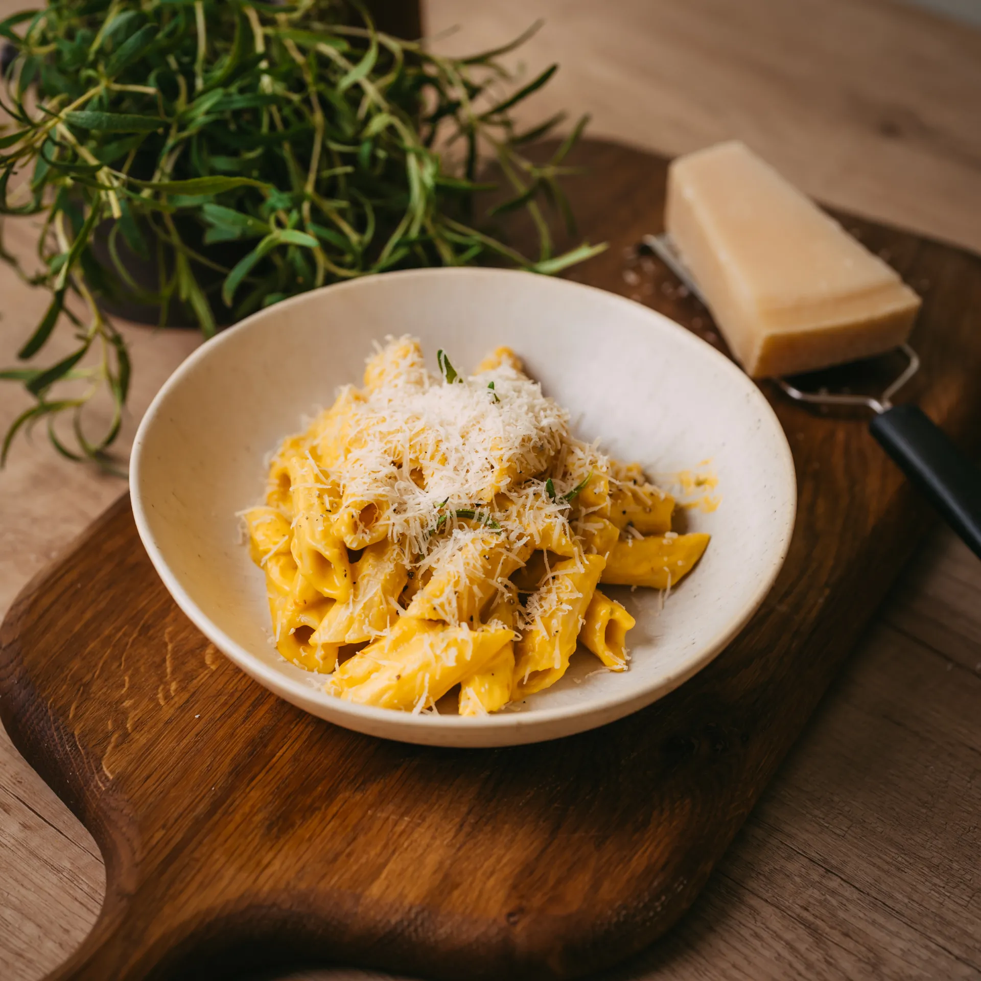 Ēdiena Fotogrāfija — A white ceramic bowl of creamy yellow rigatoni topped with grated cheese sits on a dark wooden serving board. A wedge of hard cheese rests on a grater beside fresh green herbs, all shot in warm natural light in a styled food commercial still life.