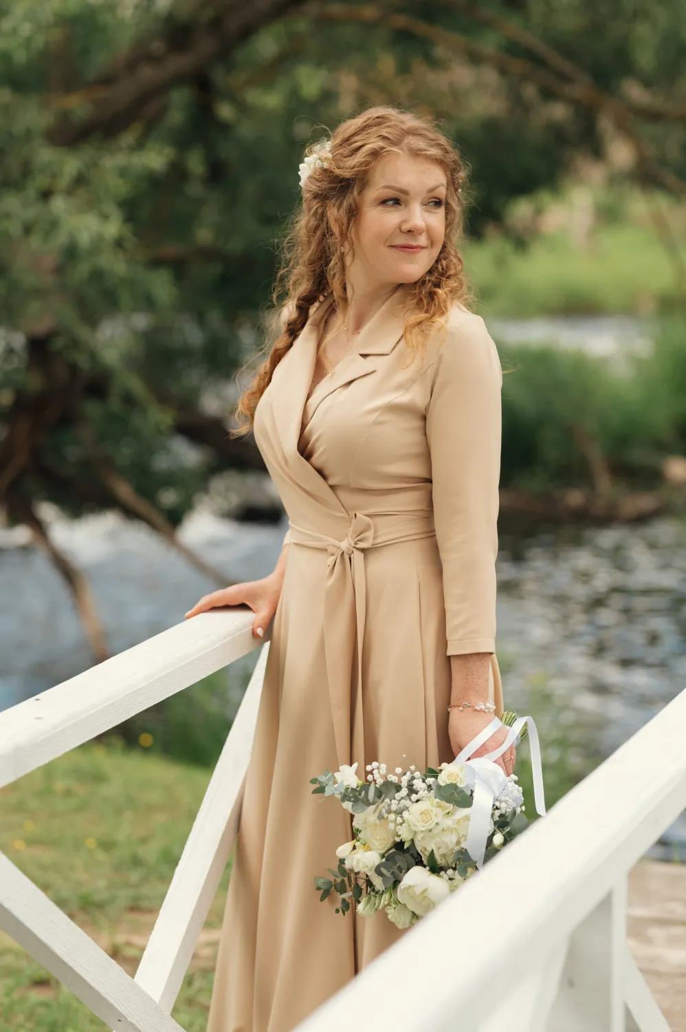 A woman with curly reddish hair adorned with a white flower stands by a white wooden bridge railing outdoors near a river and green trees. She wears a beige wrap maxi dress with three-quarter sleeves and holds a bouquet of white roses and greenery, looking over her shoulder in soft natural light.