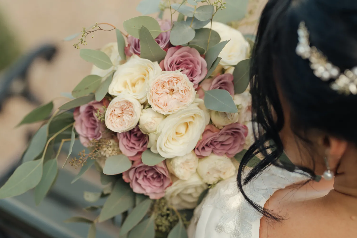 A bride in a white dress holds a bouquet of blush, cream, and mauve roses with eucalyptus leaves, viewed from behind with a jeweled hairpiece visible.