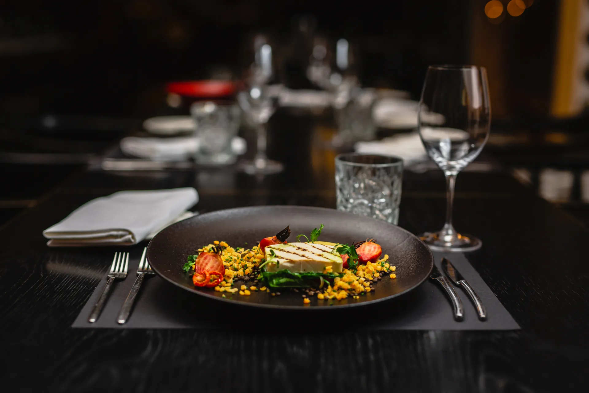 Ēdiena Fotogrāfija — A plated dish with grilled cheese, tomatoes, greens and a yellow crumble sits on a dark plate at a restaurant table. Cutlery, a folded napkin, a water glass and a wine glass are arranged on the black tablecloth, with softly blurred place settings behind in warm ambient light.