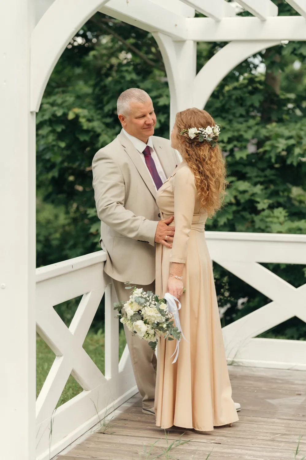 A man in a light beige suit with a maroon tie embraces a woman in a long beige dress with a white floral hairpiece from behind on a wooden deck under a white arbor. She holds a white and green bouquet; lush green foliage is behind them in a soft, natural wedding portrait style.