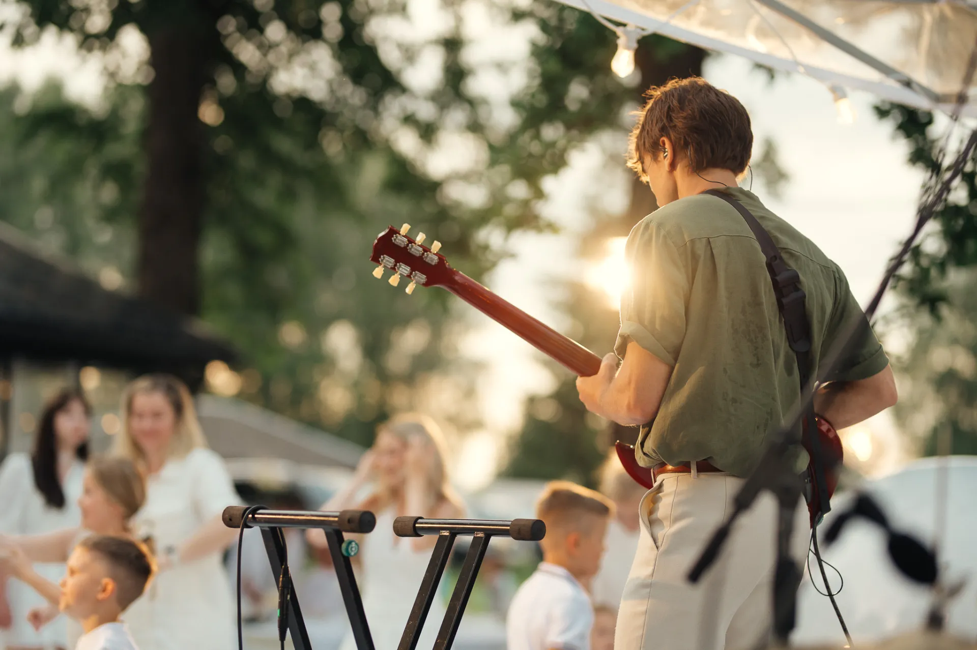 A musician in an olive shirt and light pants plays a red electric guitar on an outdoor stage at golden hour, backlit by the sun. In the softly blurred foreground and background, people in white gather, including children and women, under trees and string lights, creating a warm live-music event atmosphere.