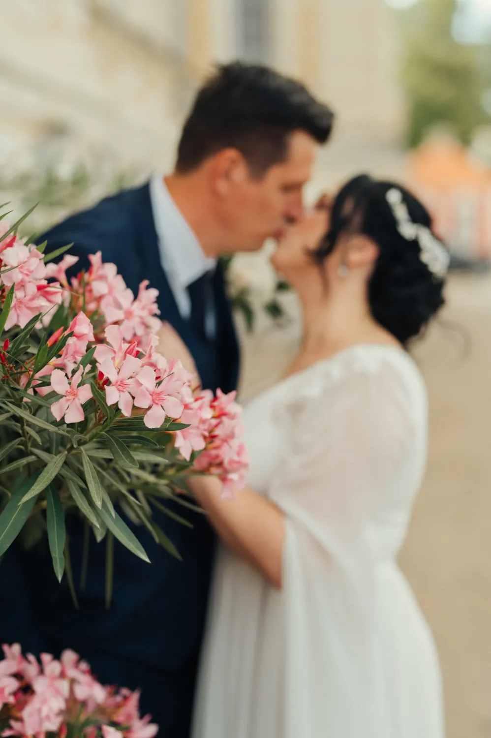 A bride in a white dress with sheer sleeves and a groom in a dark suit share a kiss outdoors, framed by pink oleander flowers in the foreground. Soft natural light creates a romantic, intimate wedding portrait with shallow depth of field.
