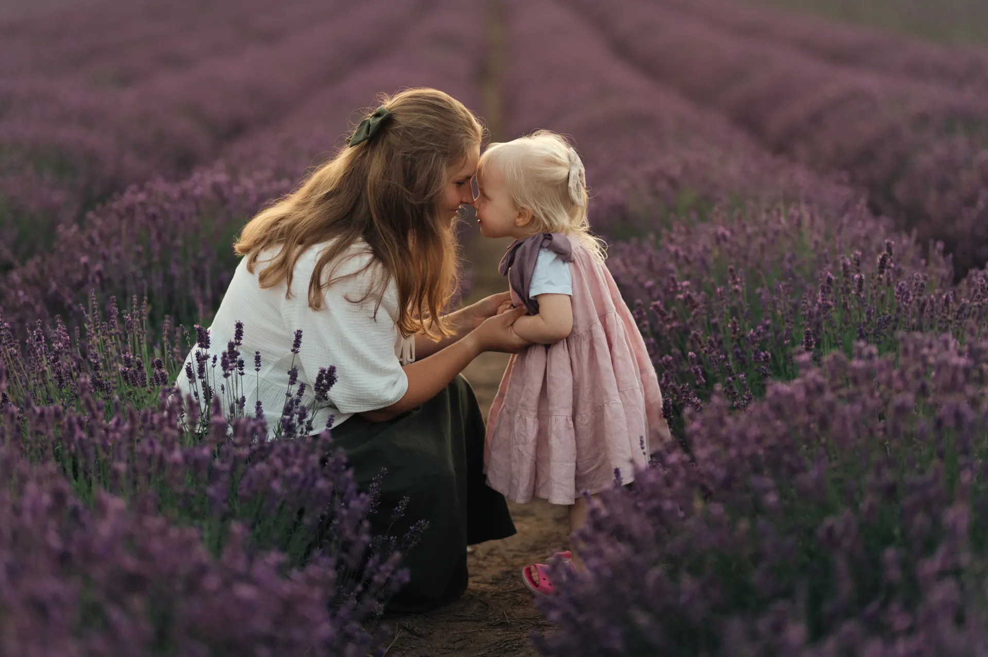 A woman with long light brown hair in a white blouse and dark skirt crouches in a lavender field at golden hour, leaning in to kiss a blonde toddler in a pink dress. The child faces her holding her hands amid rows of purple lavender. Soft warm light creates an intimate tender mood, lifestyle portrait style.