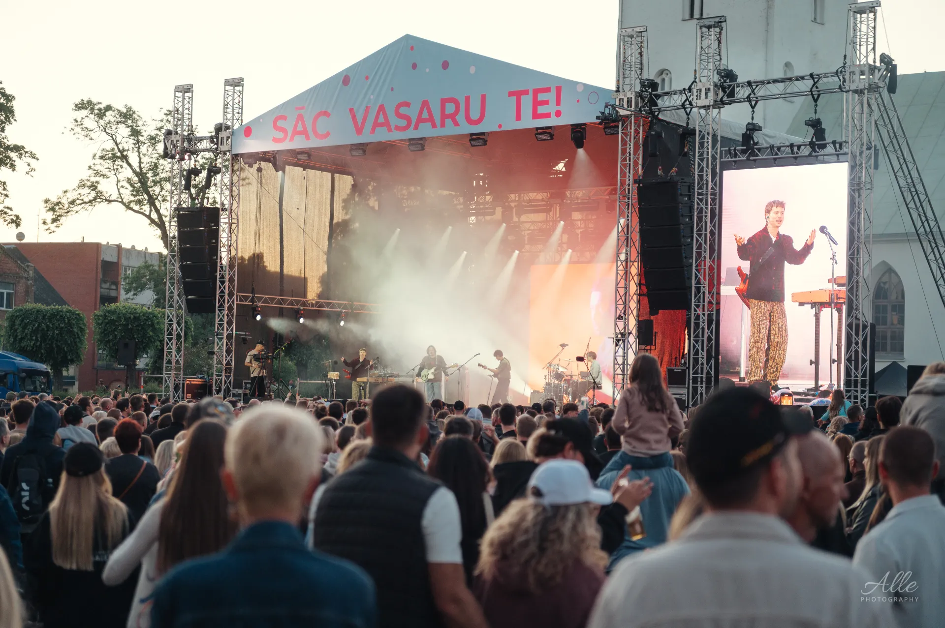 A live music concert takes place on an outdoor stage under a white tent with pink lettering, with a band performing before a large crowd and a screen showing a close-up of the singer.
