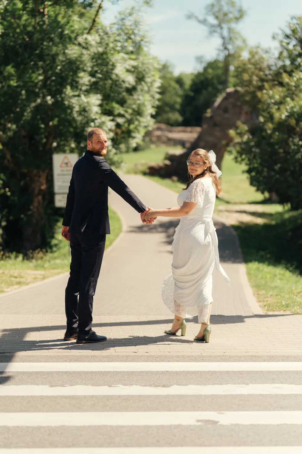 A couple holds hands on a paved path surrounded by trees, the man in a dark suit and the woman in a white lace dress with heels, captured in bright daylight with a warm, romantic mood.