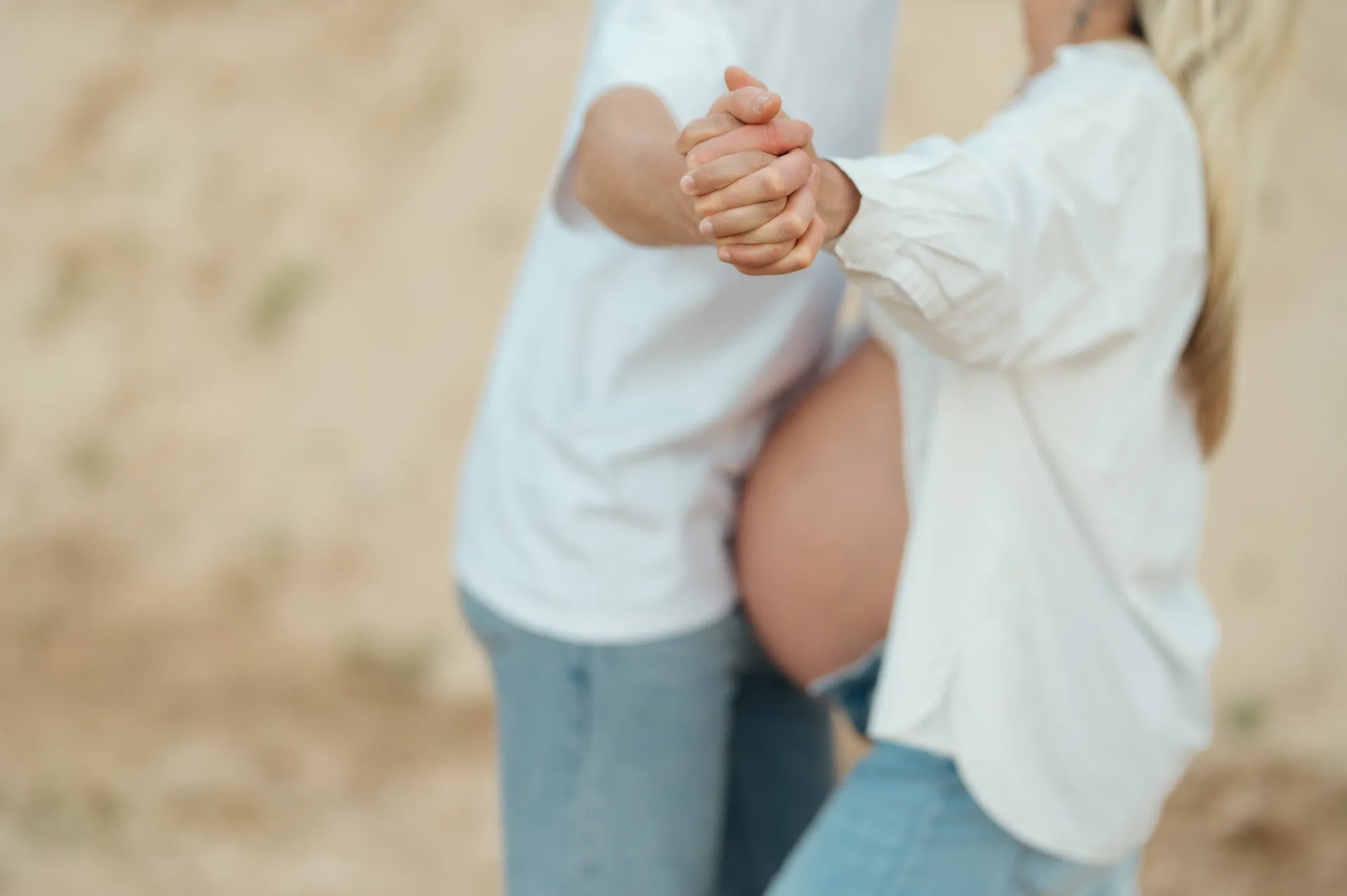 A pregnant couple holds hands outdoors against a soft, sandy background, both wearing white shirts and jeans in a tender maternity portrait.