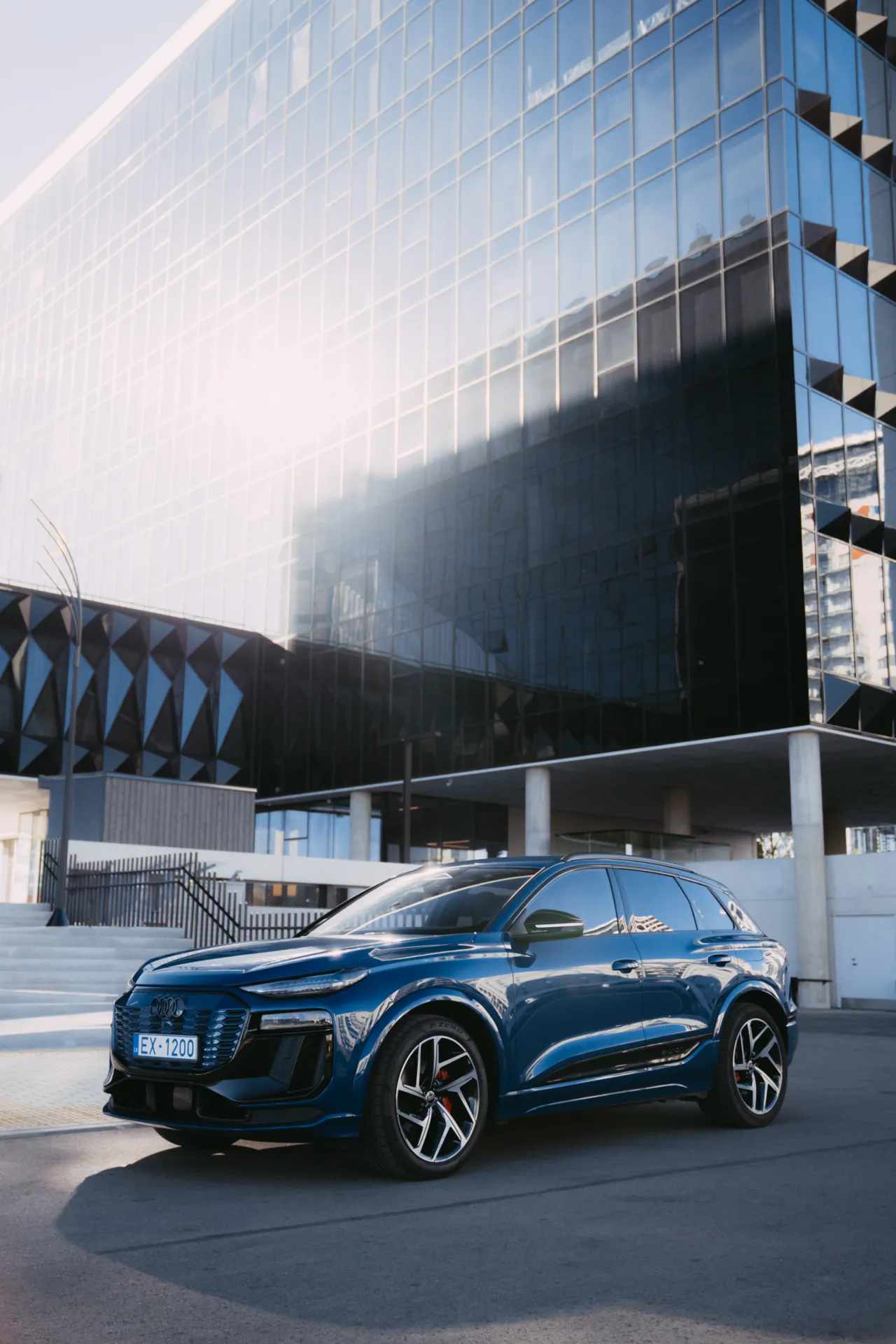 Auto Foto — A glossy blue SUV is parked on asphalt in front of a modern glass office building with bright sun glare on the facade. The commercial automotive shot shows sharp detail, reflections and strong daylight for a clean, high-end mood.