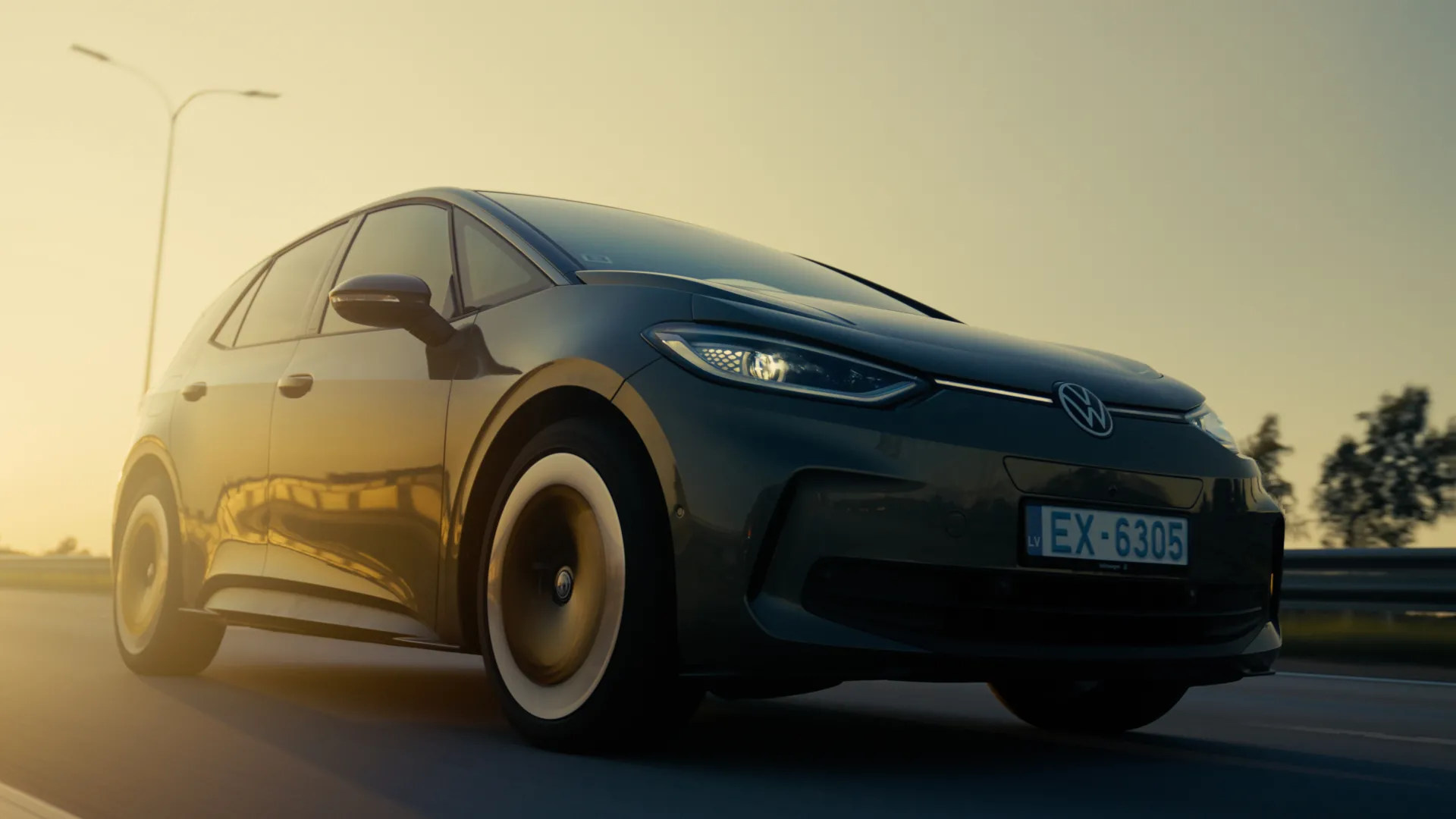 Auto Foto — A dark hatchback car with a visible VW emblem and license plate EX-6305 drives on a road at golden hour. The low-angle shot captures warm sunset light reflecting off the glossy body, with a streetlamp, trees and guardrail in the softly blurred background. Automotive commercial style.