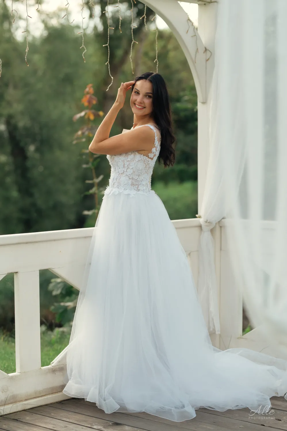 A bride in a white lace gown smiles while standing on a wooden porch, her hand raised to her hair beneath a draped arch with hanging lights, set against a soft green background.