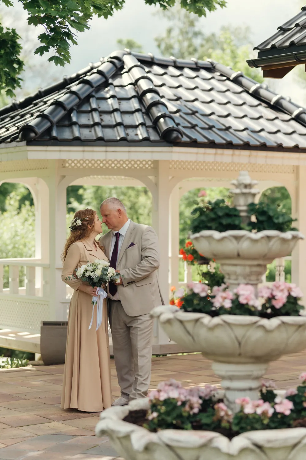 A wedding portrait of a couple embracing under a white gazebo with a tiled roof, outdoors in bright daylight. The woman in a long beige dress holds a white bouquet with a floral crown, the man in a light suit with a dark tie stands close, with a stone fountain with pink flowers in the foreground and greenery behind.