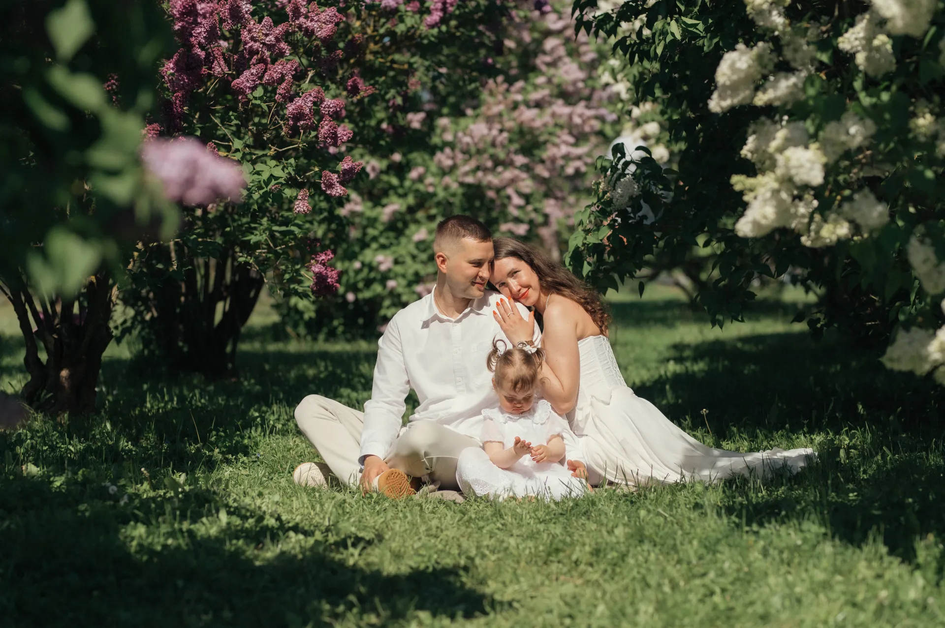 A family portrait shows a man, woman, and small child sitting together on grass beneath blooming lilac bushes, dressed in white and sharing an intimate moment in soft, dappled sunlight.