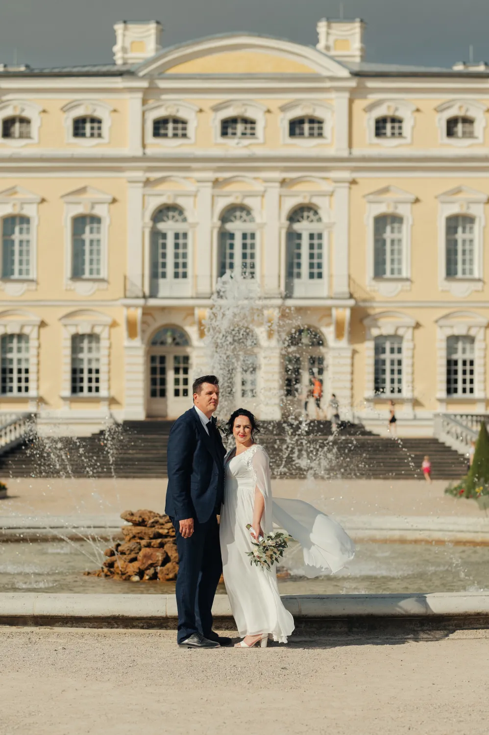 A bride in a white wedding dress holding a bouquet stands beside a groom in a dark suit in front of a large yellow palace with a fountain spraying behind them. The outdoor wedding portrait is bright and elegant with the grand building and stone steps as the backdrop.