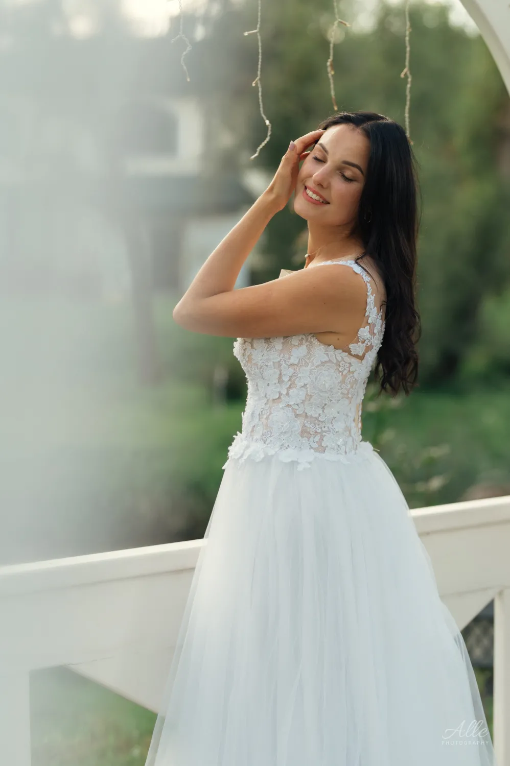 A woman in a white lace bodice wedding dress with a tulle skirt stands by a white railing outdoors, eyes closed and smiling with one hand near her face. Soft natural light and blurred greenery create a romantic wedding portrait mood.