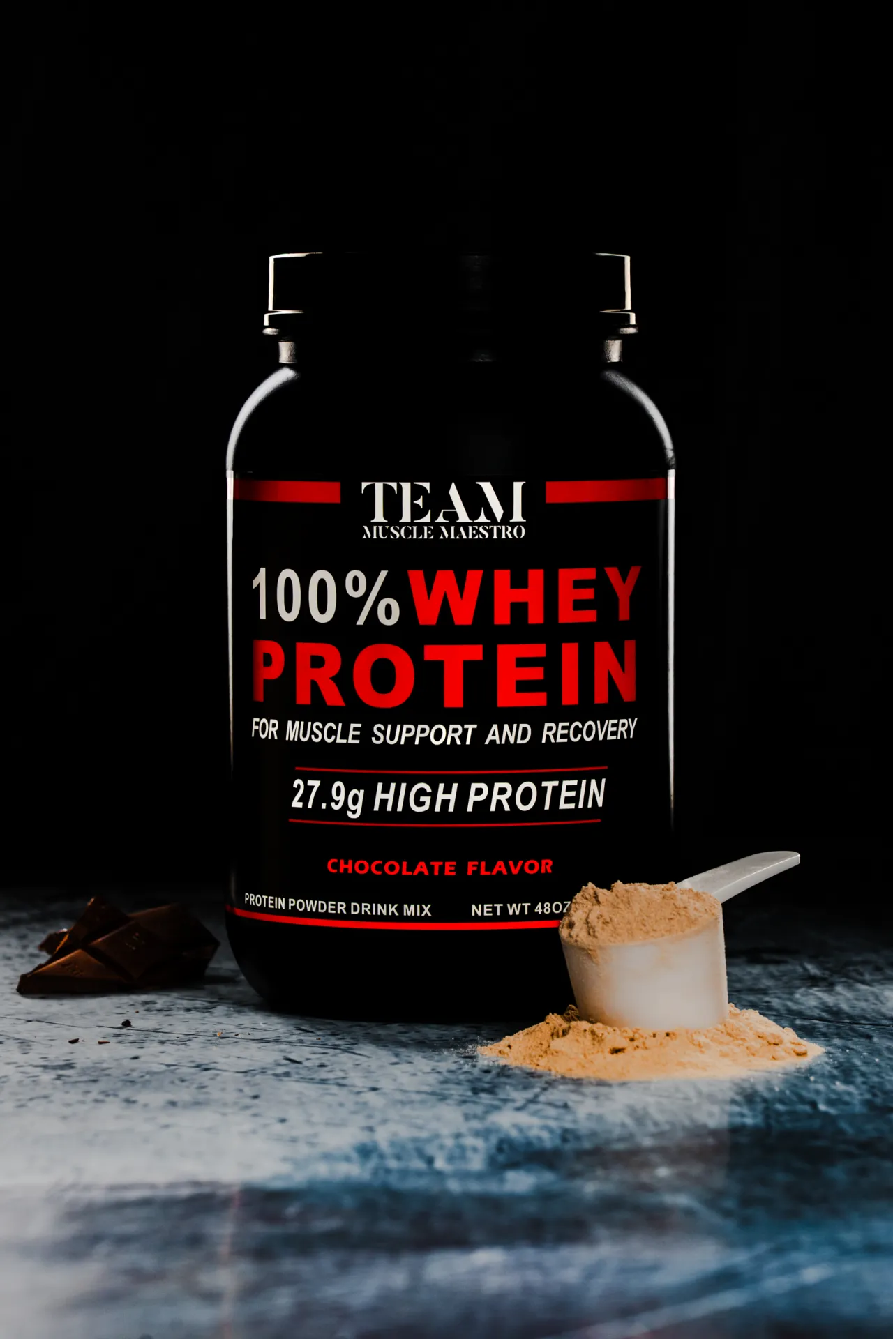 Protein powder by Muscle Maestro - Chocolate flavor — A black whey protein tub labeled 100% Whey Protein, Chocolate Flavor, sits on a textured surface with a scoop of light brown powder spilling beside it and chocolate pieces to the left, lit dramatically against a dark background in a commercial p…