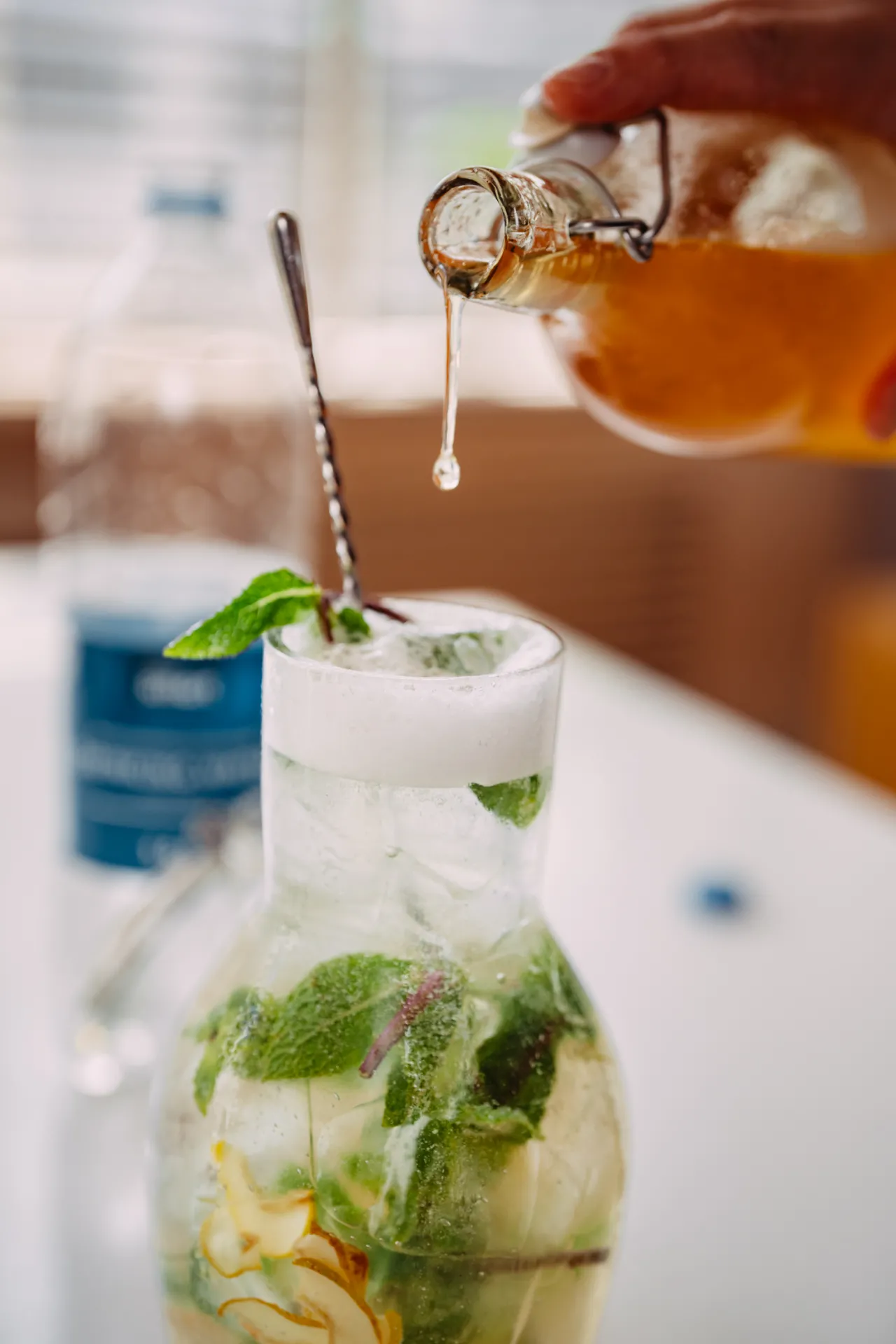 Ēdiena Fotogrāfija — A hand pours amber liquid from a glass swing-top bottle into a tall glass filled with ice, mint, and citrus slices, topped with white foam and a metal straw, in a bright commercial food and drink style.