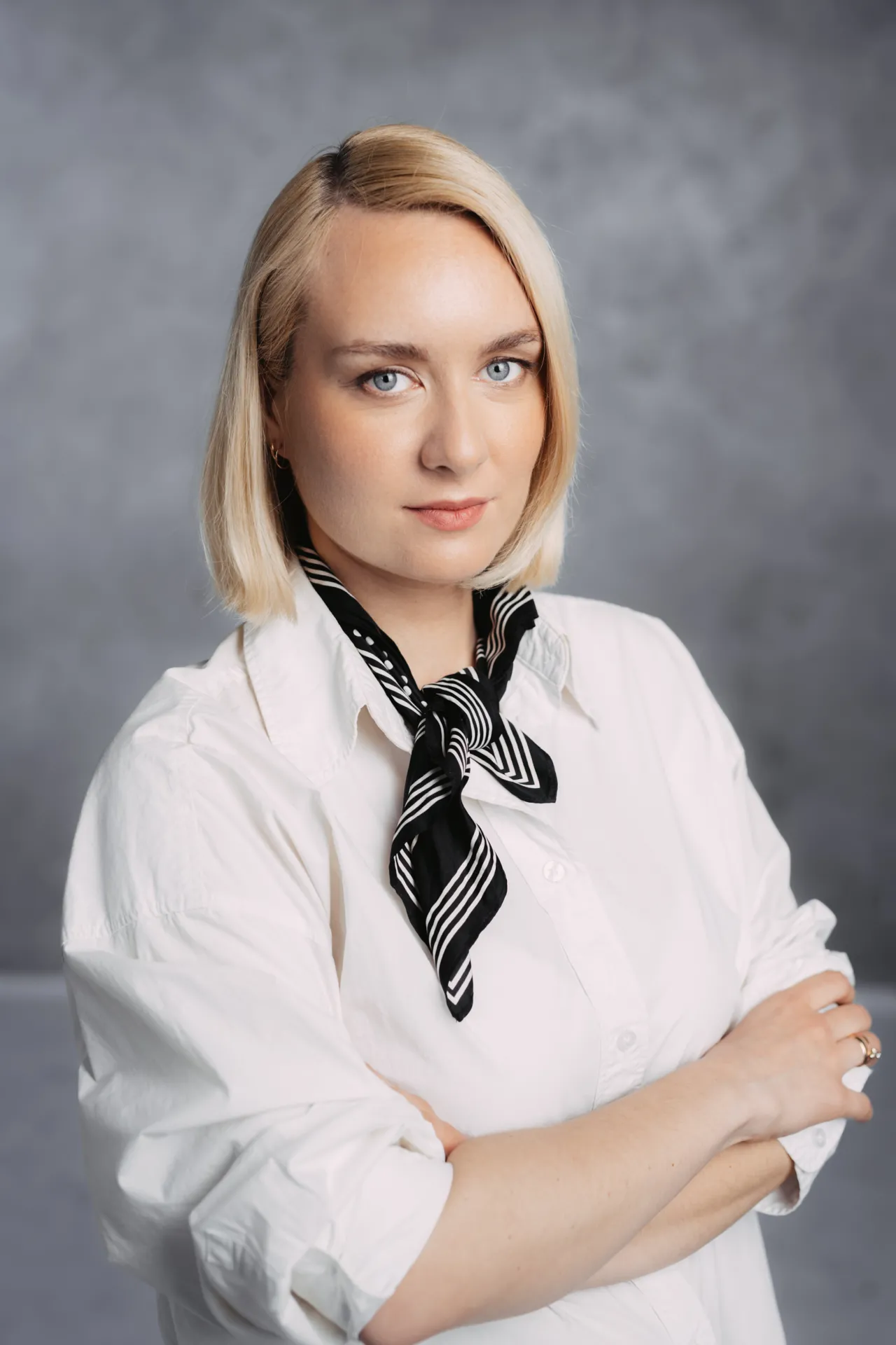 Biznesa Portreti — A studio portrait of a blonde woman with a bob haircut wearing a white button-up shirt and a black-and-white patterned neck scarf, standing against a mottled gray backdrop with her arms crossed. The lighting is soft and even, giving a calm, professional mood typical of a commerci…