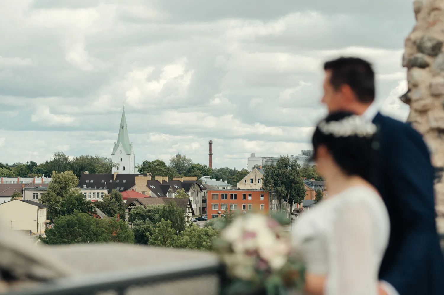 A bride and groom stand on a stone balcony overlooking a city with a church spire under a cloudy sky, captured in a soft-focus wedding portrait.