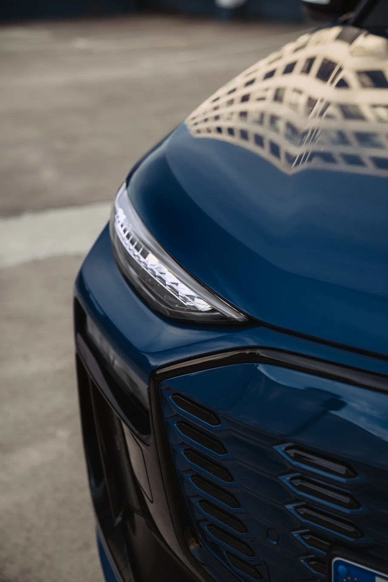Auto Foto — A close-up commercial shot of a glossy blue car's front corner, showing a sleek LED headlight and black grille with a building reflected on the hood.