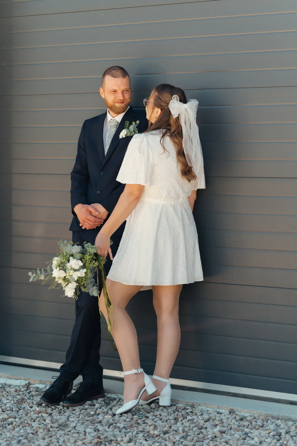 A bride in a white short lace dress with a veil bow and white heels holds a white and green bouquet while facing a groom in a dark suit with a boutonnière. They stand close together against a gray horizontally paneled wall on gravel in warm natural light, an intimate wedding portrait.