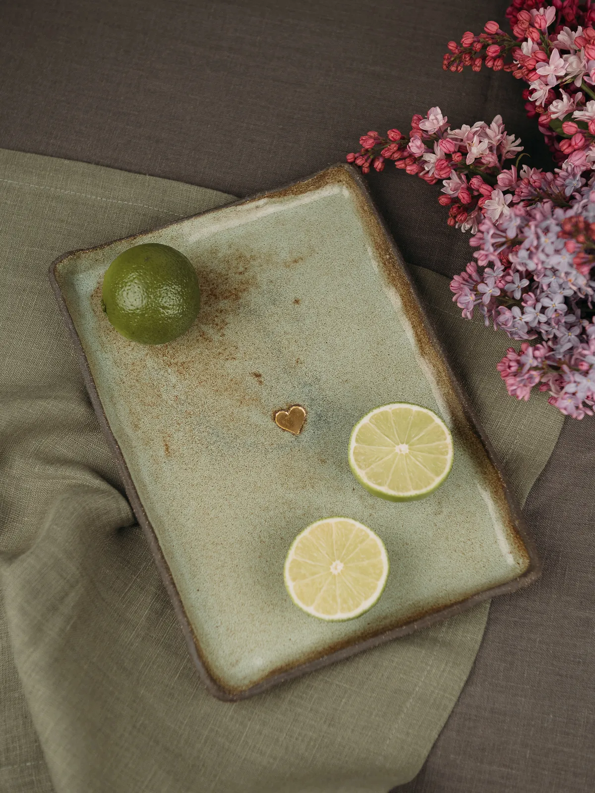 A still life composition shows a whole lime and two lime slices arranged on a rectangular ceramic tray with a small gold heart, set on a linen cloth beside pink lilac flowers under soft natural light.
