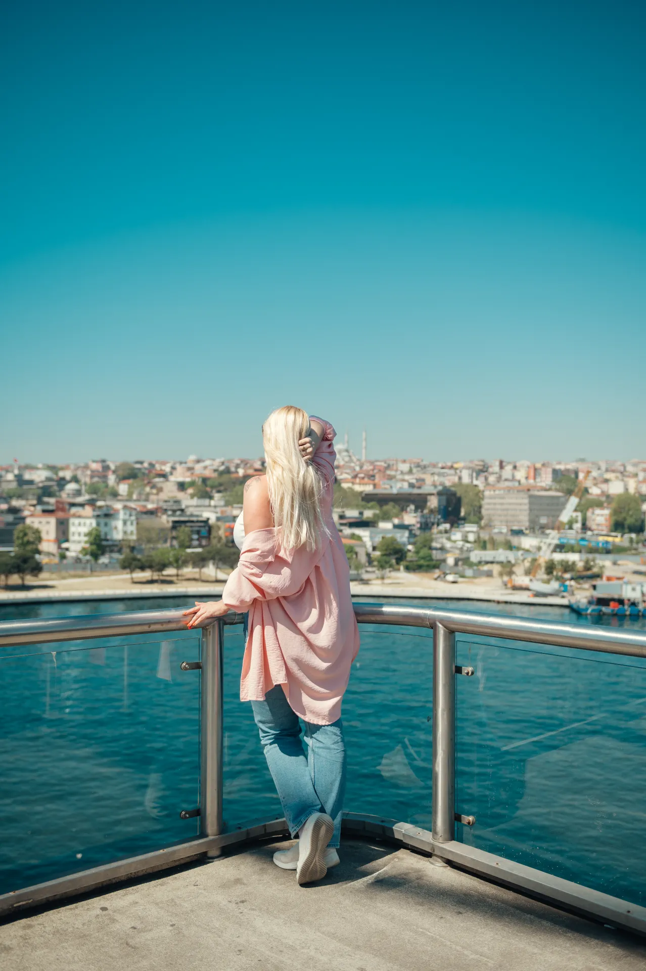 Stambula/Kapadokija — A woman with long blonde hair seen from behind leaning on a metal and glass railing overlooking bright blue water and a dense cityscape under a clear turquoise sky. She wears an oversized pink off-shoulder shirt, light blue jeans and white sneakers, with one hand in her hair.