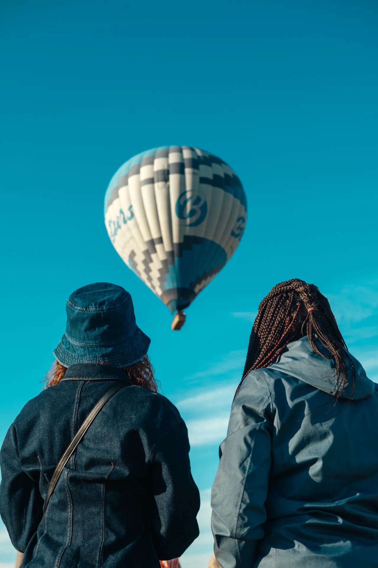 Stambula/Kapadokija — Two people seen from behind looking up at a patterned white and blue hot air balloon against a clear turquoise sky. One wears a denim bucket hat and jacket with a crossbody strap; the other has long braids and a gray hooded jacket. Bright daylight creates a calm travel lifesty…