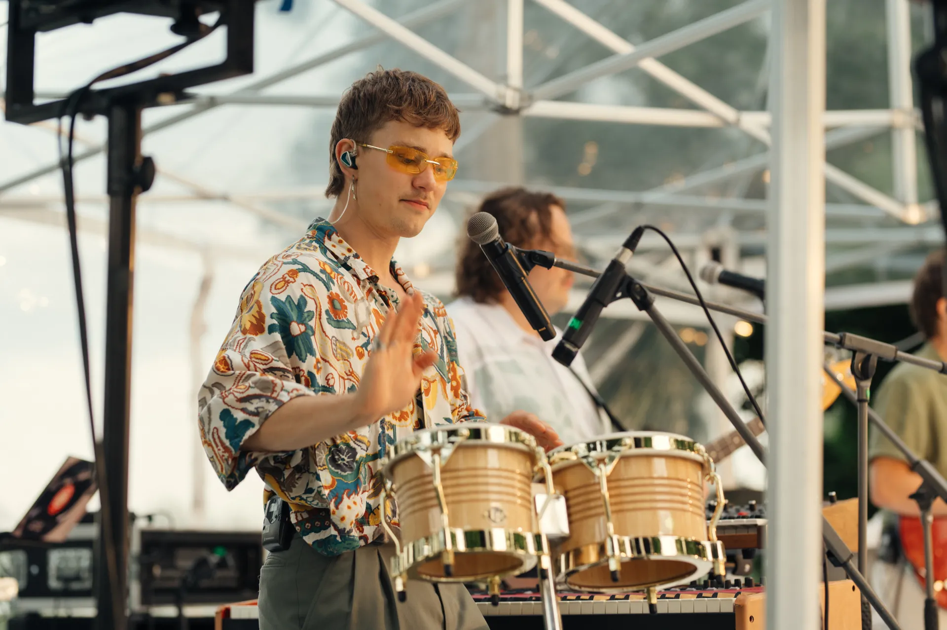 A musician wearing yellow-tinted sunglasses and a colorful floral shirt plays conga drums on stage, with microphones positioned nearby and another band member visible in the background under a white tent structure.