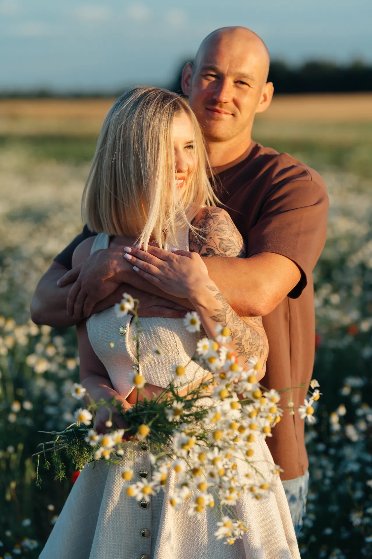 A couple embraces in a field of white daisies under warm golden light. The woman holds a large bouquet of daisies while the man stands behind her, both smiling softly in a romantic portrait.