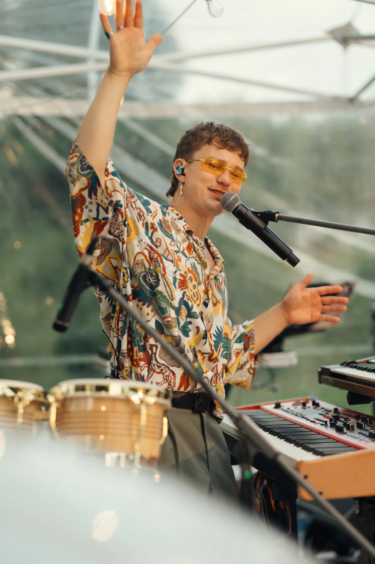 A young male musician in a colorful patterned shirt and yellow-tinted sunglasses sings into a microphone on stage, one arm raised in greeting. In the foreground are a keyboard, timbales and microphone stands under a translucent canopy, capturing a lively live performance moment with bright natural light.