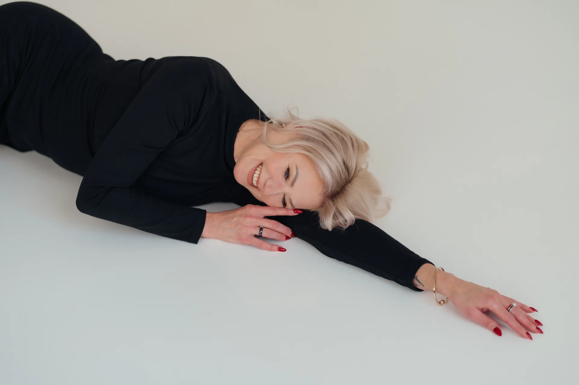 A woman with blonde hair lies on her side on a white floor, wearing a black long-sleeve dress and smiling at the camera with red nail polish visible on her hands.
