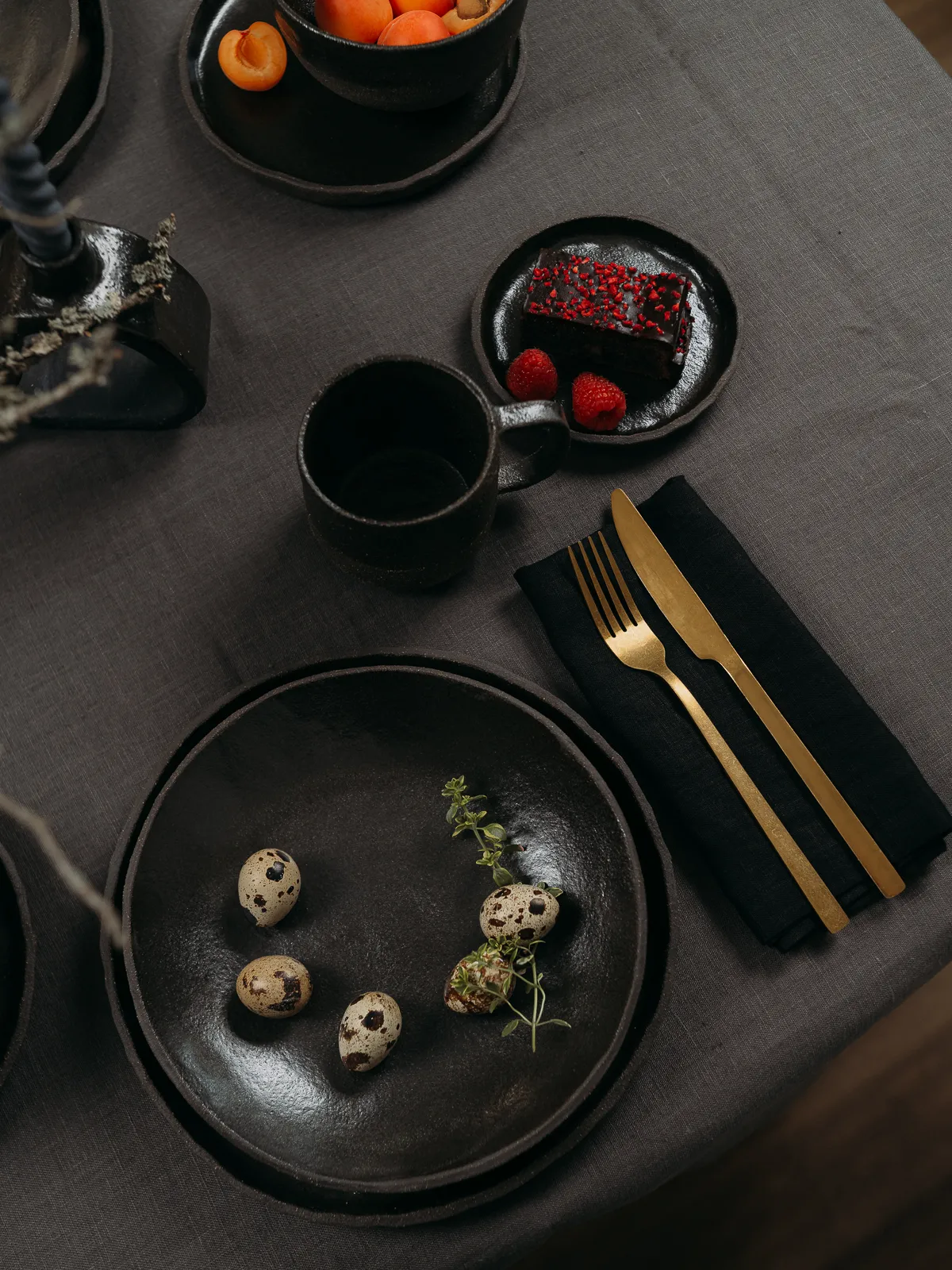 Overhead still life of a dark table setting with matte black ceramic plates, a mug, quail eggs with thyme, a chocolate dessert with raspberries, and apricots in a bowl on a gray linen tablecloth. Gold cutlery rests on a black napkin, creating a moody, minimalist food styling composition.