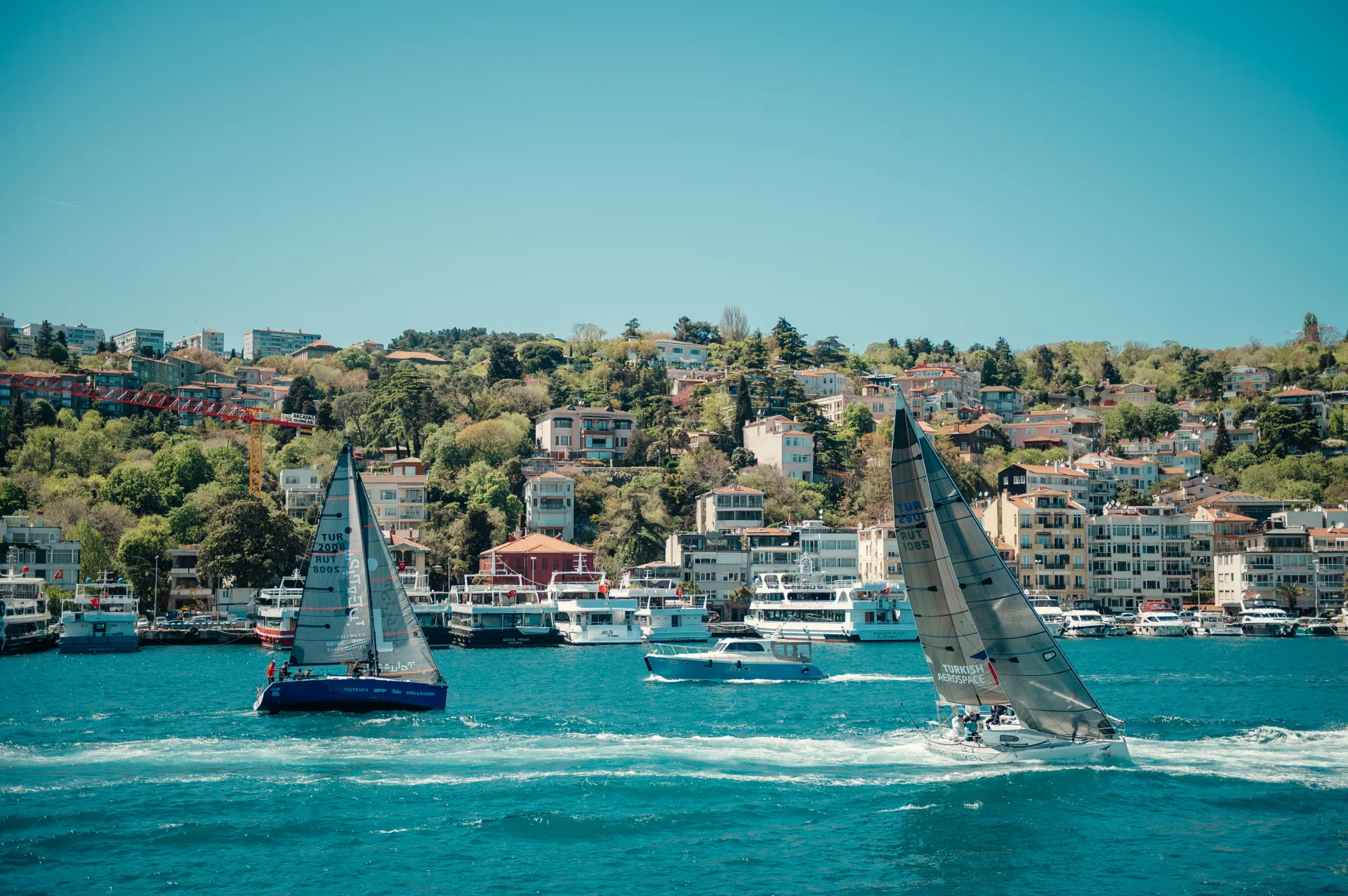 Stambula/Kapadokija — Two sailboats heel on bright turquoise water in the foreground with a small motorboat between them, docked tour boats along a shoreline backed by a densely built hillside of houses and trees under a clear blue sky. The scene captures a sunny, active nautical sports moment with…