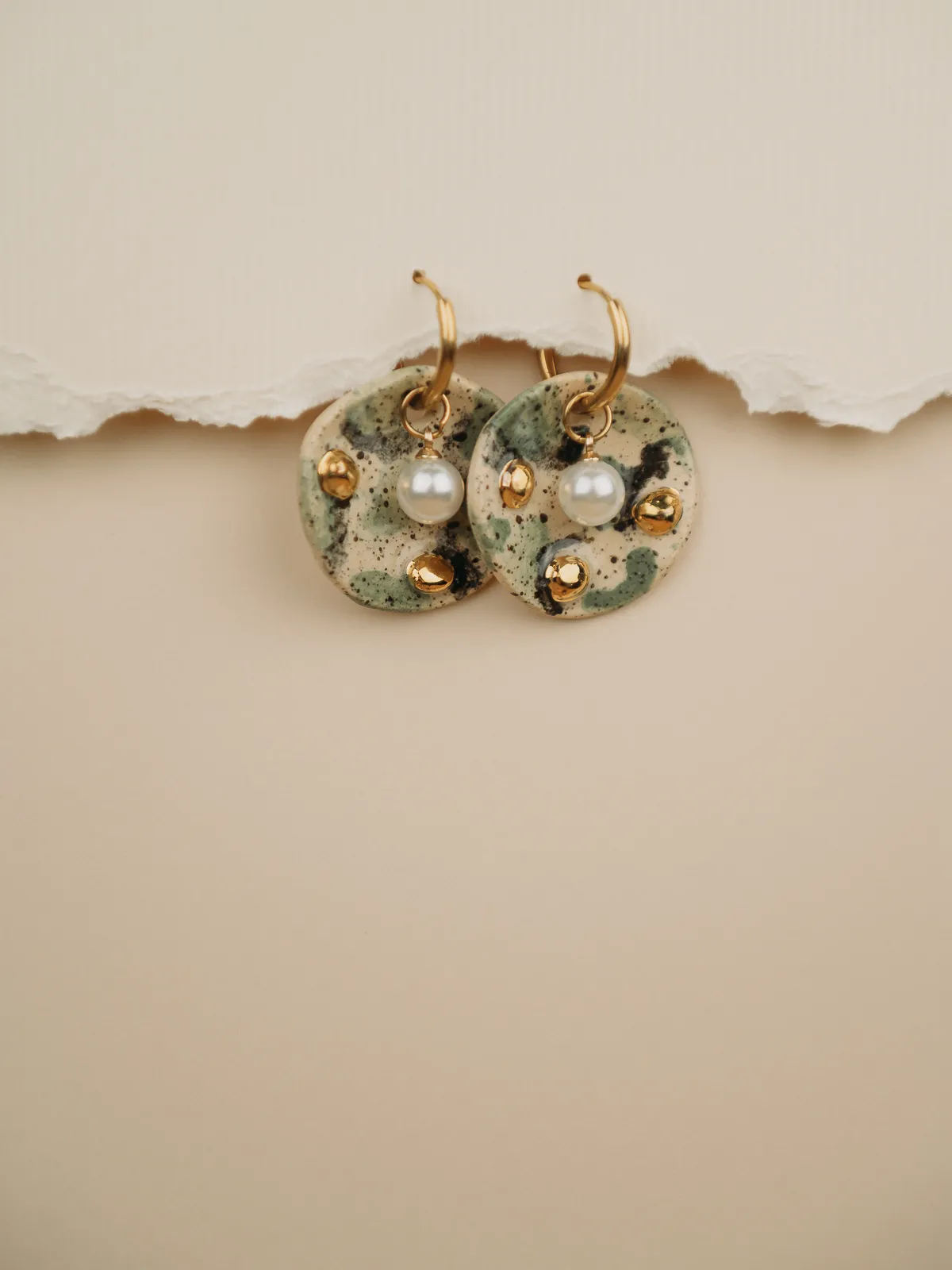 A pair of ceramic earrings with round speckled discs, gold hoops, small gold studs, and dangling white pearls rest on torn white paper against a soft beige background. Product photography with soft, even lighting.