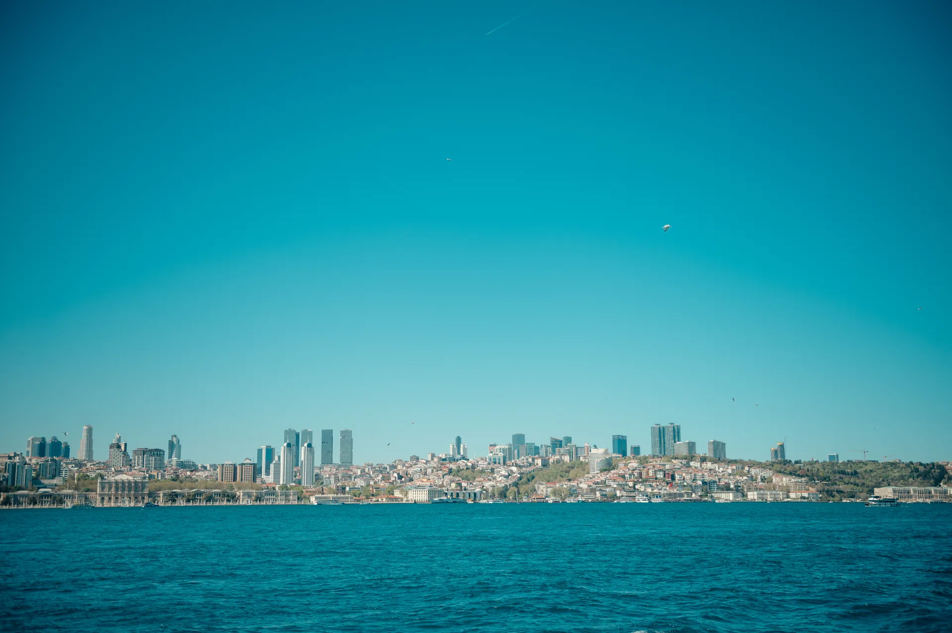 Stambula/Kapadokija — A wide city skyline stretches along a waterfront under a clear blue sky, with modern high-rises rising above dense residential buildings and calm blue water in the foreground.