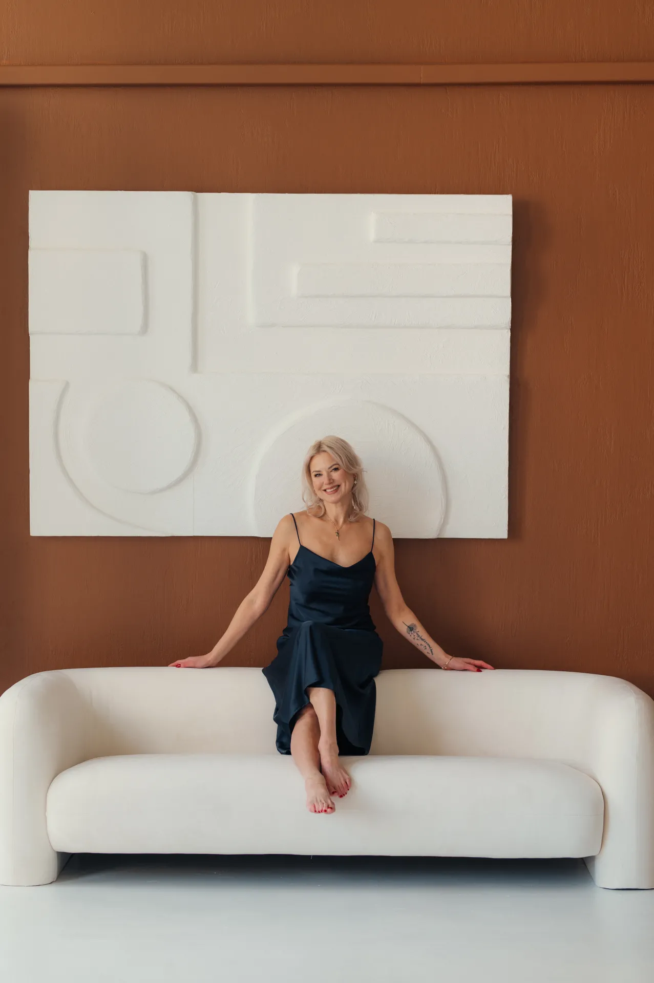 A smiling blonde woman sits barefoot on a white curved sofa in front of a warm brown wall with a large white geometric relief. She wears a dark satin slip dress, rests her hands on the sofa arms, and poses for a studio portrait with soft, even lighting and a calm, minimal aesthetic.