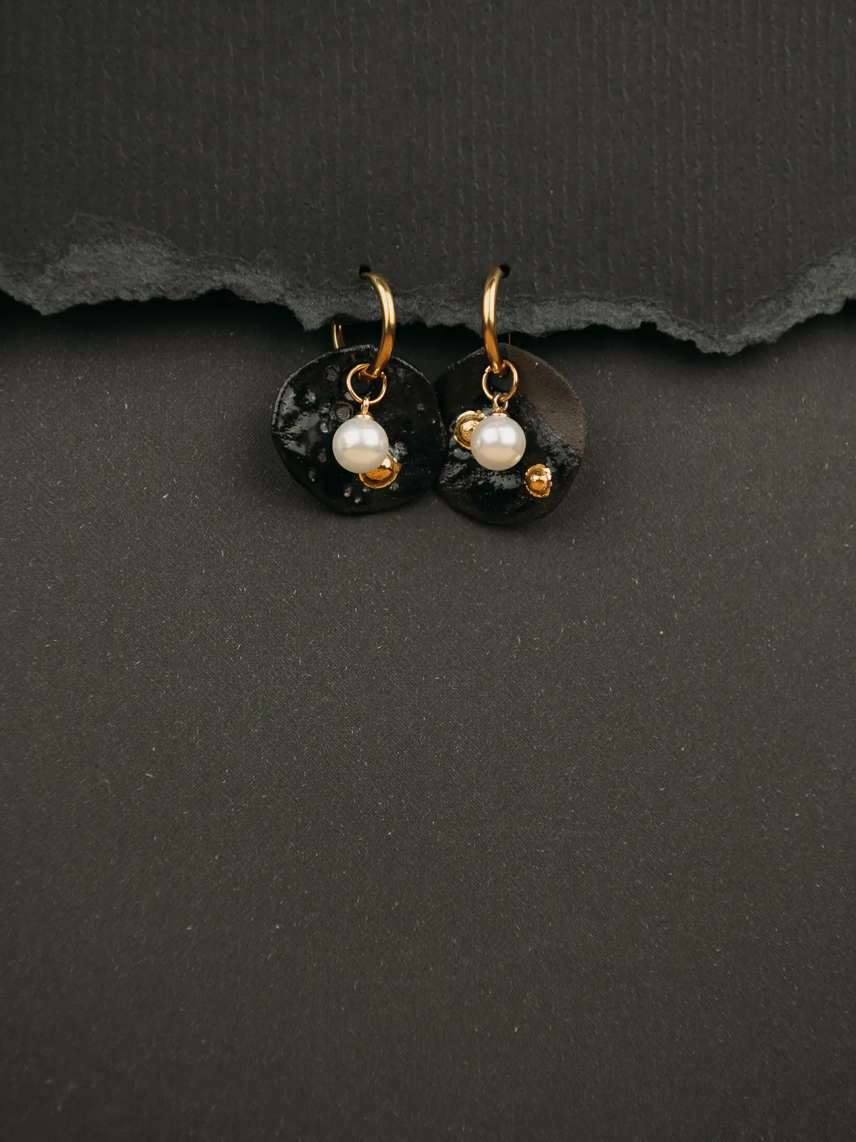 Two gold hoop earrings with black textured disc drops and small white pearl charms rest against a dark textured background with a torn paper edge above. The product-style close-up is lit softly, showing the glossy black discs, gold hardware and pearls in sharp detail against a moody dark backdrop.